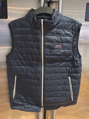 Svart dunväst från Marcoy - Snygg svart dunväst från Marcoy med quiltad design och hög krage. Västen har två dragkedjeförsedda fickor framtill och en tydlig logga på bröstet. Perfekt att slänga över en hoodie för extra värme. Stängs med silverfärgad dragkedja.