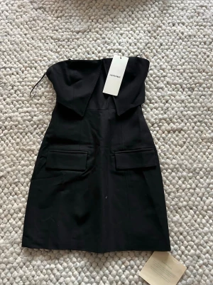 Svart axelbandslös klänning Adoore - Stilren svart axelbandslös klänning från Adoore med två stora fickor framtill, Aldrig använd 