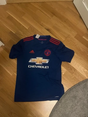 Manchester United bortatröja Pogba - Snygg mörkblå Manchester United bortatröja från Adidas med röda detaljer och Pogba 6 på ryggen. Tröjan har klubbmärke, Adidas-logga och Chevrolet som sponsor framtill. Perfekt för fotbollsfans som vill sticka ut på match eller träning.