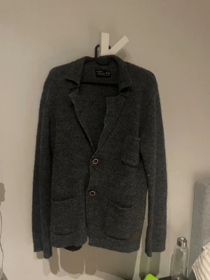 Mörkgrå stickad cardiganjacka - Mörkgrå stickad cardiganjacka med två knappar framtill, tre fickor och dekorativa knappar vid ärmsluten. Jackan har klassisk krage och lång ärm, perfekt för lager på lager och en chill stil. Materialet är mjukt och ger en avslappnad känsla.