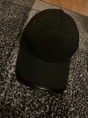 Svart keps från Fendi med logomönster - Snygg svart keps från Fendi med diskret logomönster över hela kepsen. Klassisk böjd skärm och justerbar baksida. Tillverkad i ett slitstarkt material som känns lyxigt och stilrent. Perfekt accessoar för dig som gillar exklusiva detaljer.