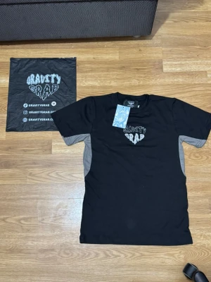 Svart Gravity Grab t-shirt med tryck, storlek S-M - Snygg svart t-shirt från Gravity Grab med vitt tryck på bröstet och gråa paneler på sidorna och ärmarna. Klassisk passform och rund hals. Perfekt för dig som gillar streetwear och vill ha en unik look, storlek S-M