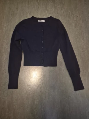 Mörkblå cropped kofta - Snygg mörkblå cropped kofta från Ivyrevel med rund halsringning och knappar hela vägen fram. Koftan har långa ärmar och ribbad midja som ger en cool siluett. Perfekt att styla över en topp eller klänning. Ganska liten i stoleken