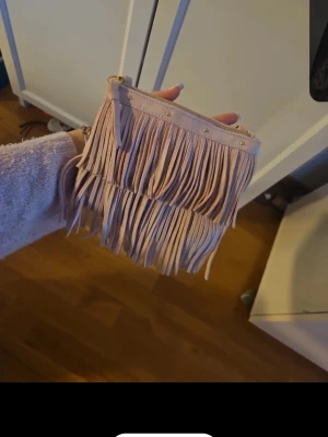 Ljusrosa axelväska med fransar - Ljusrosa axelväska med långa fransar och guldfärgade detaljer. Väskan har en justerbar axelrem och dragkedja upptill. Perfekt för dig som gillar boho-stilen och vill ha något unikt till din outfit.