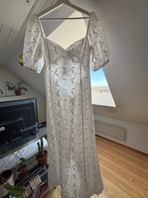 Adoore Vercelli - Romantisk vit långklänning med blommigt mönster och puffiga kortärmar. Klänningen har v-ringad front och knytning i ryggen för extra detalj. Perfekt för sommarens alla tillfällen och ger en drömmig look.