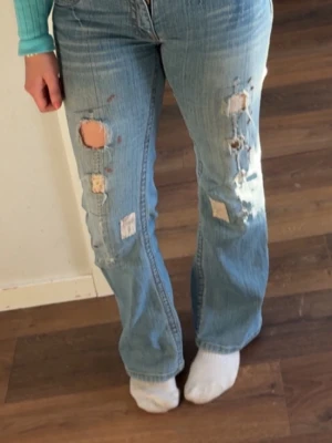 Ljusblå bootcut jeans med patchar - Säljer ett par ljusblå bootcut jeans med hål samt patchningar på för de ej passar i storleken längre.         Jeansen är vintage och köpta i Paris på second hand, samt med skräddarsytt tyg på vilket ger dig en väldigt unik look. Storlek eller märke stod ej på jeansen vid köp, men uppmäter att de är ca 36/38. Perfekt för dig som vill ha en modern och alldeles egen style! Midjemått finns med på bild 4, och önskas det fler bilder så är det bara att höra av sig!💕
