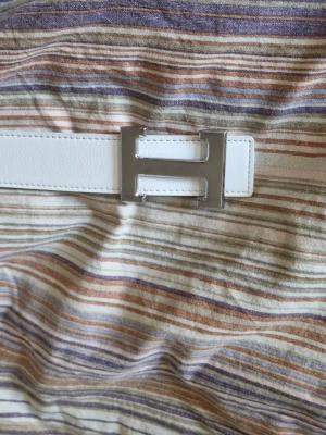 Vitt skärp med silverfärgat H-spänne - (Helt nytt och oanvänt) Snyggt vitt skärp med ett stort, stilrent H-format spänne i silverfärg. Skärpet har en clean look och är gjort i ett slätt material som ger en exklusiv känsla. Perfekt för att lyfta vilken outfit som helst.