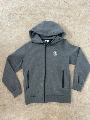 Grå hoodie från Moncler - Snygg grå hoodie från Moncler med dragkedja och två svarta fickor framtill. Hoodien har ett klassiskt Moncler-märke på bröstet och är tillverkad i mjukt bomullsmaterial. Perfekt för dig som gillar stilrena och exklusiva plagg.
