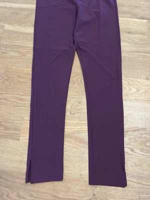 Lila leggings med slits vid benslut - Snygga lila leggings med raka ben och diskret slits vid benslut. Materialet är stretchigt och mjukt, perfekt för en chill och trendig look. Passar dig som gillar att sticka ut med färg och enkel design.