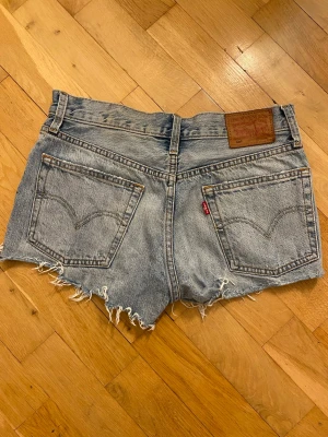 Levi's ljusa denimshorts med fransar - Säljer ett par ljusa denimshorts från Levi's med råa, fransiga kanter och klassiska femficksdetaljer. Shortsens midja är hög och de har en vintage look med slitningar och en brun läderpatch bak. Perfekta för sommarens alla äventyr.
