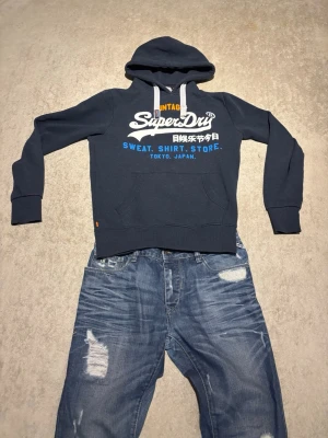 Mörkblå hoodie från Superdry - Mörkblå hoodie från Superdry med stor vit och blå logga samt japansk text på bröstet. Klassisk känguruficka framtill och justerbar huva med vita snören. Tillverkad i mjukt bomullsmaterial, perfekt för en avslappnad streetwear-look.