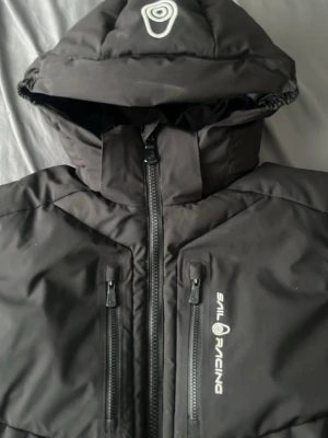 Svart dunjacka från Sail Racing - Säljer en svart dunjacka från Sail Racing med huva och två dragkedjeförsedda bröstfickor. Jackan har en stilren design med logga på bröstet och huvan. Perfekt för kalla vinterdagar och riktigt varm tack vare dunfyllningen.