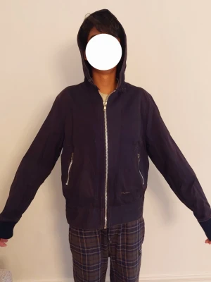 Svartgrå tunn jacka  med huva från Elvine - Gråsvart jacka från Elvine med huva och dragkedja framtill. Två snedställda fickor med dragkedja och ribbade muddar vid ärmslut och midja. Storlek L men känns som M pga kort modell