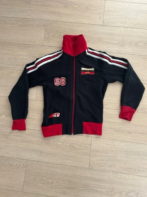 Svart och röd ziptröja med tryck - Sportig ziptröja i svart med röda detaljer och vita ränder på ärmarna. Tröjan har hög krage, muddar och tryck med siffran 68 samt patchar på bröstet och nedtill. Perfekt för en avslappnad streetwear-look.