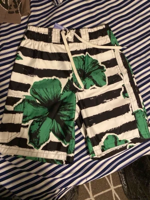 Randiga badbyxor från Ocean Surf XL - Snygga badbyxor från Ocean Surf i storlek XL. De har svartvita ränder och stora gröna hibiskusblommor. Dragsko i midjan och praktisk ficka på sidan. Tillverkade i 100% polyester, perfekta för stranden eller poolen.