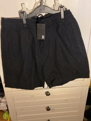 Svarta shorts från 157 i linne - Snygga svarta shorts från 157 i luftig linnemix. Modellen har elastisk midja med dragsko och två sidofickor. Perfekta för varma dagar och enkel att matcha med olika stilar.