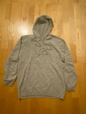 Ljusgrå kashmir hoodie - Mysig ljusgrå hoodie i mjukt stickat material. Klassisk modell med huva och dragsko med metalländar. Perfekt för chill dagar och enkel att matcha med jeans eller mjukisbyxor.