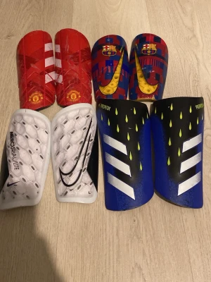 Det är 250kr styck! - Säljer ett set med sex par benskydd från Nike och Adidas. Färgerna är röd, blå, vit, svart och gul med olika klubbmotiv som Manchester United och FC Barcelona. Tillverkade i plast med vadderad insida för extra komfort. Perfekta för fotbollsträning och match.