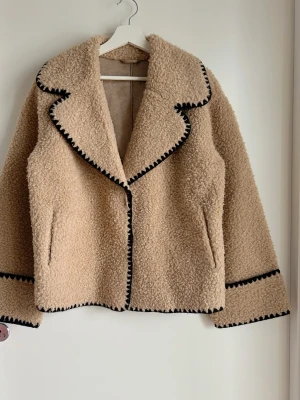 Beige teddyjacka med svarta detaljer - Mysig beige teddyjacka med markerade svarta sömmar längs krage, ärmslut och kanter. Jackan har bred krage och två fickor framtill. Perfekt för dig som vill ha en trendig och varm jacka med unik look. Bröstmått: 114 cm Längd: 66 cm 