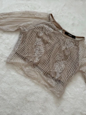 Beige spetsblus från Bik Bok - Säljer en beige spetsblus från Bik Bok. Blusen har ett transparent, blommigt spetsmönster och tunna ränder. Croppad modell som ger en trendig look. Passar som xs/s