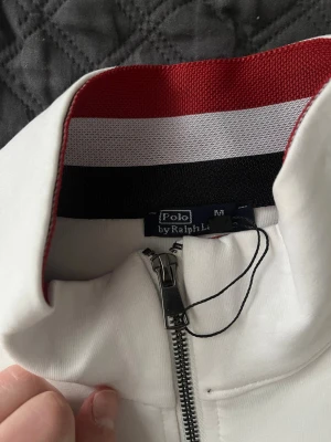 Vit ziptröja från Polo Ralph Lauren - Snygg vit långärmad ziptröja från Polo Ralph Lauren med röd logga på bröstet och halv dragkedja. Kragen har röd och vit insida för extra detalj. Perfekt till jeans eller chinos för en clean look.