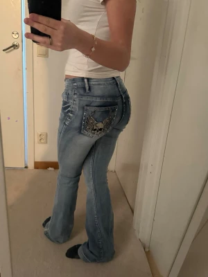 Jeans med tryck på rumpan - Säljer ett par ljusblå bootcut jeans med cool döskalle och vingar på bakfickan. Jeansen har slitningar och kontrastsömmar för en edgy look. Storleken funkar för både xs och s. Ingen aning om vart dom är köpta då de it står. Använda ungefär 2 gånger