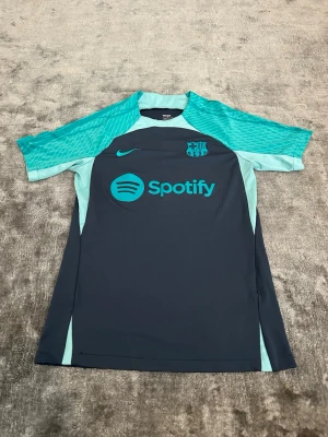 Barcelona tränings t-shirt Nike - Snygg Barcelona tränings t-shirt från Nike i mörkblått med turkosa detaljer. Tryck med Spotify-logga och klubbmärke på bröstet. Tillverkad i ett lätt och funktionellt material som passar perfekt till träning.