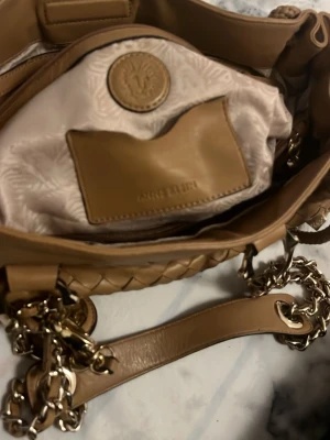 Beige handväska från Anne klein  - Snygg beige handväska från Anne klein i flätat läder med guldfärgade kedjedetaljer och logotypdetaljer. Väskan har ett innerfack med läderkant och präglad logga samt dekorativa metalldetaljer med Anne klein ikoniska lejonsymbol. Väldigt fin och aldrig använd 