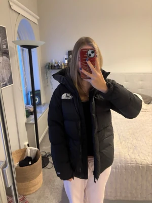 Svart pufferjacka från The North Face - Säljer en svart pufferjacka från The North Face med stor huva och logga på bröstet. Jackan har en boxig passform, dragkedja framtill och är riktigt fluffig. Perfekt för kalla dagar och ger en cool streetwear-look.