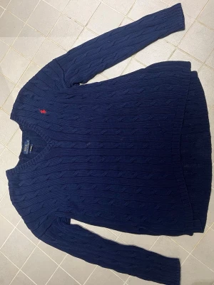 Mörkblå kabelstickad tröja Polo Ralph Lauren - Kabelstickad mörkblå tröja från Polo Ralph Lauren med klassisk röd broderad logga på bröstet. Tröjan har v-ringning, långa ärmar och ribbade muddar. Perfekt för dig som gillar tidlös och stilren design.