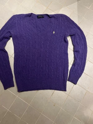 Lila kabelstickad tröja Polo Ralph Lauren - Snygg lila kabelstickad tröja från Polo Ralph Lauren med V-ringning och det klassiska broderade logot på bröstet. Tröjan har långa ärmar och ribbade muddar vid ärmslut och nederkant. Perfekt för dig som gillar stilrena och tidlösa plagg.