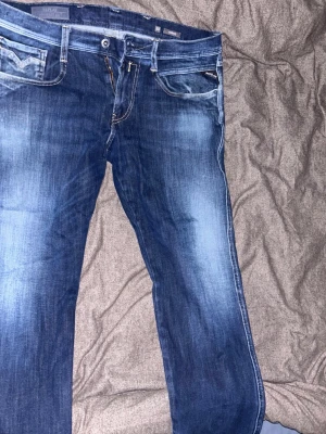 Blå jeans från Replay med slitningar - Säljer ett par snygga blå jeans från Replay med klassisk femficksmodell och coola slitningar framtill. Jeansen har raka ben och detaljerade sömmar på bakfickorna. Perfekta för dig som gillar en avslappnad men trendig stil. 33/32