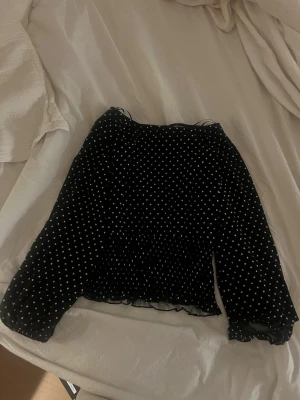 Svart prickig blus med volang - Supersöt svart blus med vita prickar och volangdetaljer. Blusen har långa ärmar med lätt puff och resår vid ärmslutet. Tunn och luftig, perfekt för att styla med jeans eller kjol. Ger en trendig och lekfull look! Liten hål vid ena ärmen men man ser inte det eller märker de 