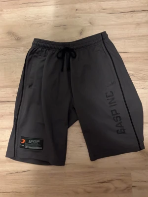 Svarta träningsshorts från GASP - Snygga svarta träningsshorts från GASP med tryck på ena benet och siffran 89 på baksidan. Shortsen har dragsko i midjan och är tillverkade i ett lätt och ventilerande syntetmaterial. Perfekta för gymmet eller andra sportaktiviteter.