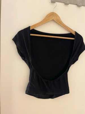 Svart croppad topp med uppen rygg  - Snygg svart croppad topp med öppen rygg  och bred urringning bak. Kort ärm och figurnära passform, perfekt till höga jeans eller kjol. Tillverkad i mjukt stretchigt material för en bekväm känsla.