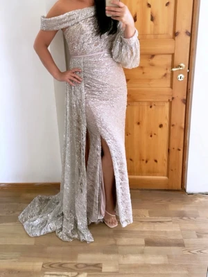 Glittrig silverfärgad långklänning - Säljer en elegant, glittrig långklänning i silver med oneshoulder-design och slits framtill. Klänningen har en draperad detalj över axeln och ett vackert fallande tyg. Perfekt för dig som vill sticka ut på bal eller fest. Det medföljer även påsen/skyddet för klänningen. De enda slitaget är botten som har blivit smutsig av marken. 