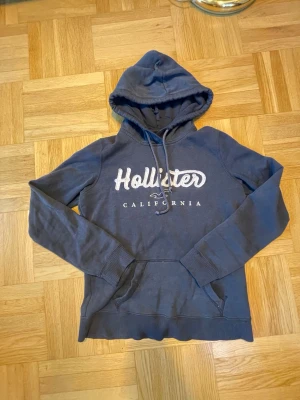 Mörkblå Hollister hoodie  - Snygg mörkblå hoodie från Hollister med vit broderad logga och texten 'California' på bröstet. Klassisk känguruficka framtill, dragsko i huvan och ribbade muddar vid ärmar och nederkant. Perfekt för en avslappnad och cool stil. Bra skick men andvänd.