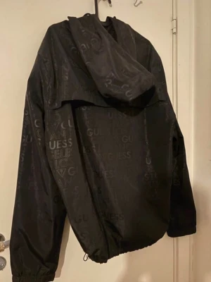 Svart vindjacka från Guess - Snygg svart vindjacka från Guess med heltryckt logotypmönster och dragkedja framtill. Jackan har huva, resår vid ärmslut och nederkant samt meshfoder på insidan för extra komfort. Perfekt för dig som vill ha en stilren och trendig look.