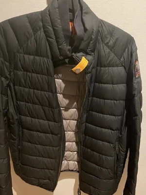 Svart dunjacka från Parajumpers - Snygg svart dunjacka från Parajumpers med gul detalj vid kragen och logotyp på ärmen. Jackan har quiltad design, hög krage och dragkedja framtill samt praktiska sidofickor. Perfekt för kalla vinterdagar och riktigt stilren look.