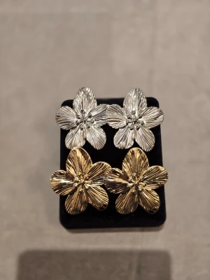Blommiga örhängen i guld och silver - Säljer två par stora örhängen i form av blommor, ett par i blankt silverfärgat metall och ett par i guldton. Örhängena har tydliga ådringar i metallen som ger en cool struktur och en riktigt statement-look. Perfekta för dig som vill sticka ut med accessoarerna!