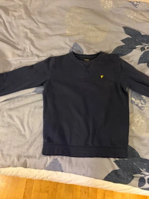 Mörkblå sweatshirt från Lyle & Scott - Mörkblå tröja från Lyle & Scott med klassisk rund halsringning och gul logga broderad på bröstet. Tröjan har långa ärmar och ribbade muddar vid ärmslut och nederkant. Perfekt för en avslappnad och stilren look.