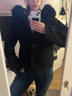 Svart kappa med fuskpäls - Stilren svart kappa med stora knappar och krage i mjuk fuskpäls. Rak modell med fickor på sidorna och dekorativ knapp vid ärmslut. Perfekt för dig som vill ha en klassisk men trendig look.