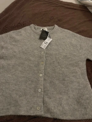 Ljusgrå kofta från Gina Tricot - Mysig ljusgrå kofta från Gina Tricot i ribbstickad ullblandning med rund hals och knappar framtill. Koftan har en avslappnad passform och är tillverkad av 74% RWS-certifierad ull. Perfekt för lager på lager och enkel att styla.