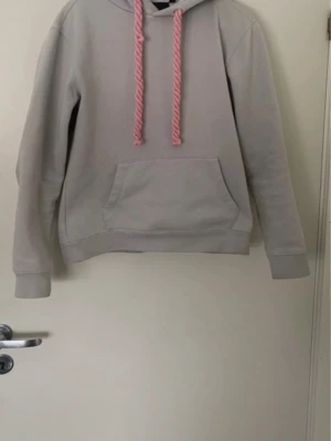 Ljusgrå hoodie med rosa snören syna world - Ljusgrå hoodie från syna world med stora rosa snören i huvan och en klassisk magficka framtill. Tillverkad i mjuk bomullsblandning för extra komfort. Perfekt för en avslappnad och trendig look.