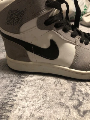 Air Jordan 1 High grå/vit/svart - Först till kvarn. Snygga Air Jordan 1 High sneakers i grått, vitt och svart. Ovandel i läder och mocka med klassisk svart swoosh och Jordan Wings-logga på sidan. Perforerad tåbox för ventilation och svarta skosnören. Perfekta för dig som gillar streetwear och ikoniska sneakers.
