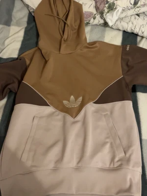 Beige och brun hoodie från Adidas - Säljer en snygg hoodie från Adidas i beige och bruna nyanser med stor huva och magficka. Ikoniska Adidas-loggan framtill och diskret Adidas-tryck på ärmen. Hoodien har en modern färgblock-design och är gjord i mjukt material, perfekt för en avslappnad streetwear-look.