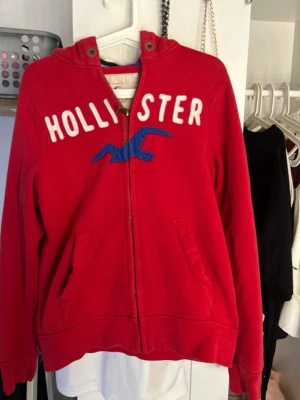 Röd hoodie från Hollister med dragkedja - Snygg röd hoodie från Hollister med vit text och blå logga på bröstet. Hoodien har dragkedja framtill, två fickor och en klassisk huva. Perfekt för en avslappnad och trendig stil. Materialet känns mjukt och bekvämt.