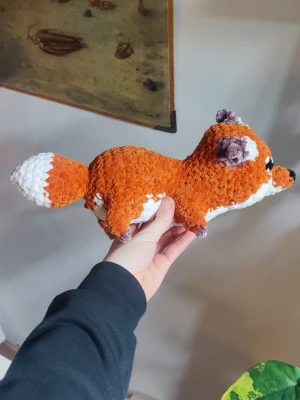 Mysig virkad räv mjukisdjur - Handvirkat mjukisdjur i form av en räv. Räven är orange med vita och bruna detaljer, svart nos och stora broderade ögon. Tillverkad i mjukt garn som gör den extra gosig och perfekt som prydnad eller leksak.