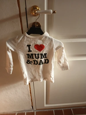 Vit långärmad topp H&M baby - Söt vit långärmad topp från H&M med trycket 'I ❤️ Mum & Dad' framtill. Tillverkad i mjuk bomull, perfekt för små barn. Passar storlek 86 (12-18 månader). Enkel och bekväm modell med rund hals.