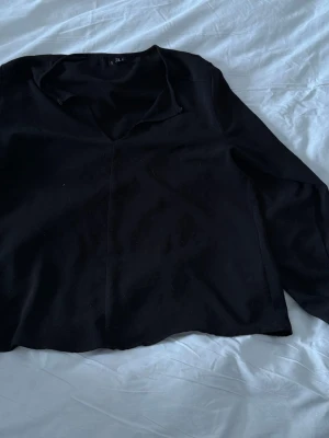 Svart blus från Vero Moda - Stilren svart blus från Vero Moda med v-ringning och krage. Blusen har långa ärmar och är tillverkad i ett mjukt, lätt tyg som ger en avslappnad men ändå chic look. Perfekt att matcha med jeans eller kjol för en trendig outfit.